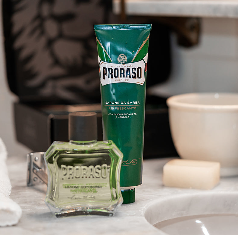 Components of Refresh Classic Shaving Duo After Shave Lotion and Shave Cream Tube sitting on a counter with the box open in the back. Other items on the white marble table include silver razor, Post-Shave Stone and white shave bowl